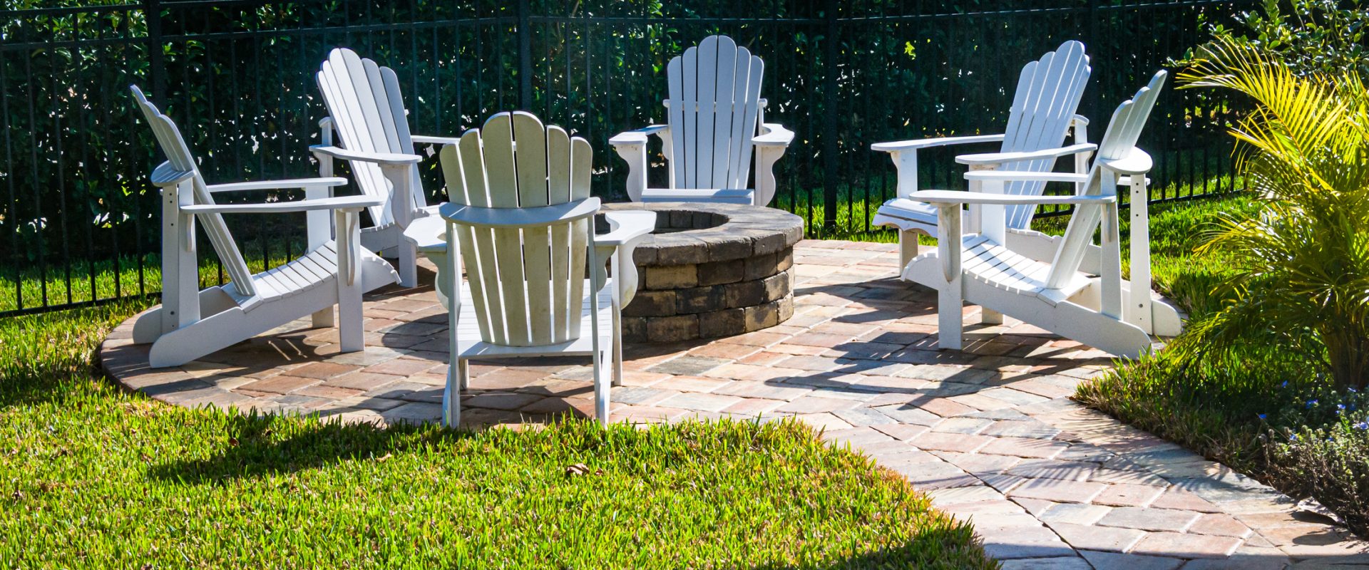 White Adirondack chairs arranged around a circular stone fire pit on a brick patio in a sunny backyard with lush hedges and a black fence behind.