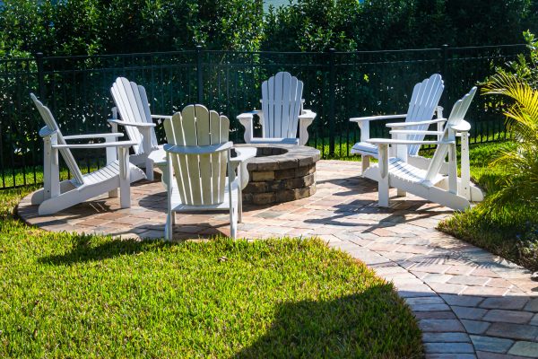 White Adirondack chairs arranged around a circular stone fire pit on a brick patio in a sunny backyard with lush hedges and a black fence behind.